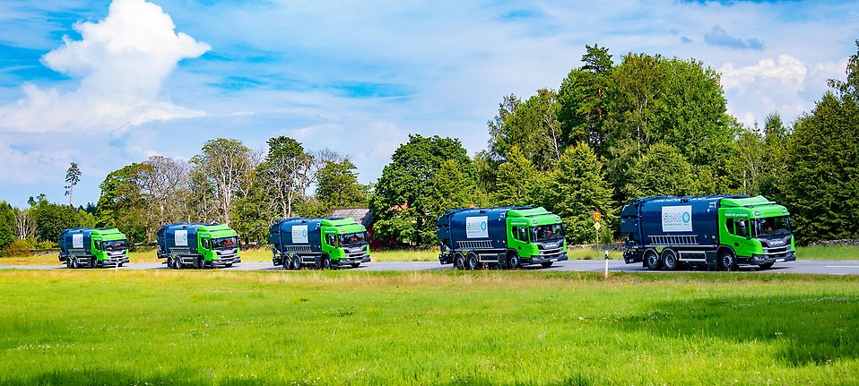 Gröna och blå sopbilar kör på rad genom ett sommarlandskap.